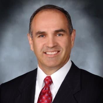Smiling man in a dark pinstripe suit and red tie against a mottled grey background.